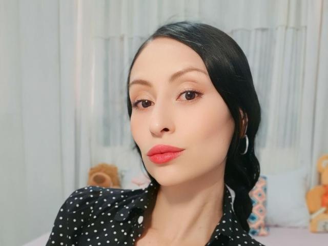 AliceRossiX has pale skin, black hair in a side braid, red lipstick, hoop earrings, and wears a black polka dot shirt. Her tits are small, and she's looking at the camera.