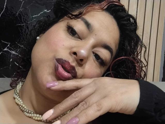 LarussoSophi, con cabello oscuro y rizado, reflejos rosados, un collar de perlas, un labial púrpura intenso y uñas rosas, frunciendo ligeramente los labios y apoyando la mano en su rostro..