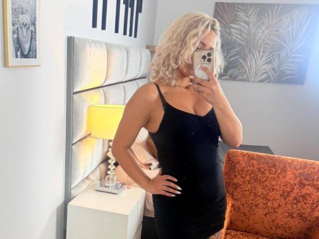 ElenaSaenz with curly blonde hair, wearing a black, tight, sleeveless dress, showing her ass, standing in a bedroom, taking a mirror selfie, holding a phone.