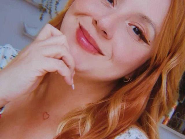 PauleetteRoux a les cheveux roux, un sourire charmant, et elle touche son menton avec son doigt. Elle a un petit tatouage en forme de cœur sur sa poitrine. Ses seins sont partiellement visibles..