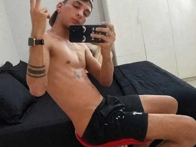 Shirtless Daom69 with a slim build, black shorts, and tattoos, sitting on a bed, making a peace sign, holding a phone, wearing a watch, with a confident expression.