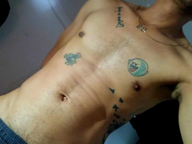 Le torse nu de Daom69, orné de tatouages, notamment un crabe, un visage de dessin animé bleu et une ligne d'arbres sur le côté droit. Il porte un pantalon sombre, dont le bas est légèrement visible..