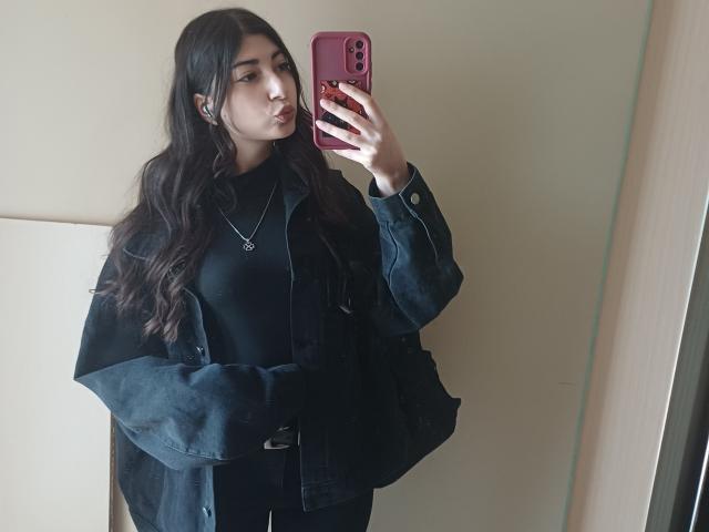 OnieAndreatta mit langen, dunklen Haaren, voller Lippen, die ein rosa Handy hält, trägt ein schwarzes Oberteil und eine Jacke und steht in einem Spiegel-Selfie, wobei sie eine kleine Halskette zeigt..