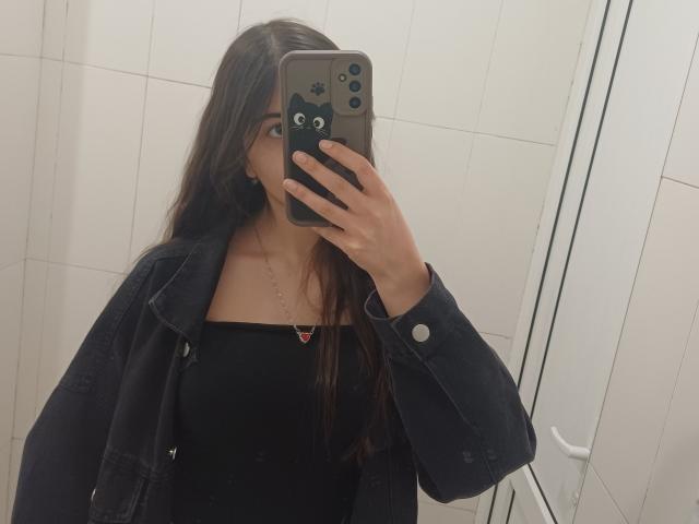 OnieAndreatta mit langen, braunen Haaren, trägt ein schwarzes Top mit Trägern, eine Jeansjacke und eine Halskette und macht ein Selfie vor einem Spiegel mit ihrem Handy, auf dem eine Katzen-Hülle zu sehen ist..