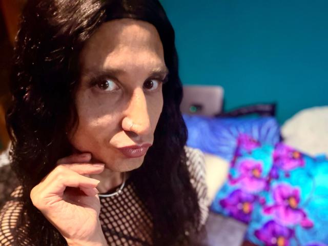 MaryXXL-hot with long black wavy hair, brown eyes, and nose ring, wearing a black fishnet top, touching her chin with her hand, looking slightly to the side.