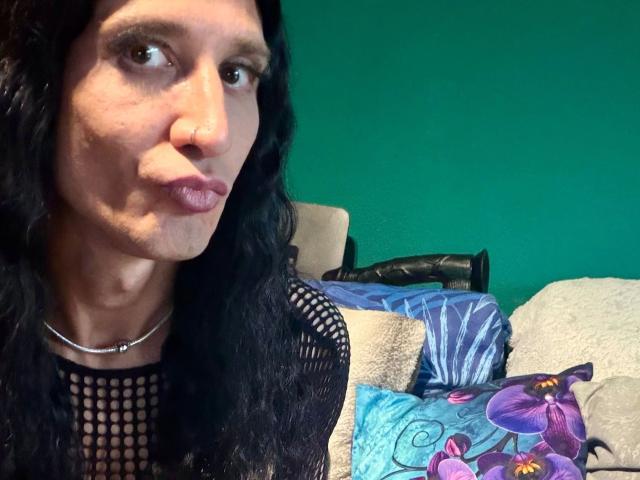 MaryXXL-hot with long black wavy hair, nose ring, and fishnet top, pouting lips, green wall, black dildo on beige couch, colorful pillows, and blue-green patterned pillow.