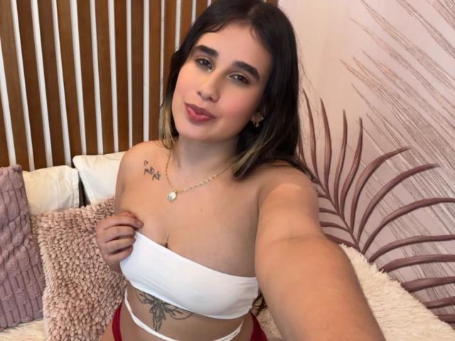 KatieDesirX, avec une poitrine de taille moyenne, tenant un haut blanc, montrant un tatouage et un tatouage floral sur son côté, souriante, cheveux bruns foncés, rouge à lèvres rose, collier en or, sous-vêtements rouges..