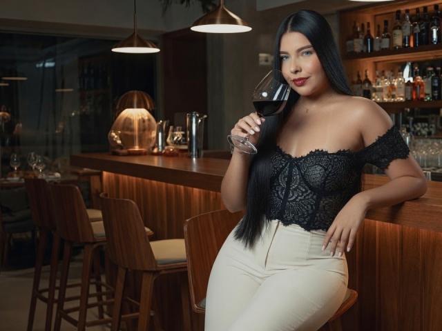 Sofia Delacruz, con lunghi capelli neri, una blusa bianca con pizzo e pantaloni bianchi, tiene in mano un bicchiere di vino rosso, ed è in piedi davanti a un bancone di legno con sgabelli. Il seno è visibile..