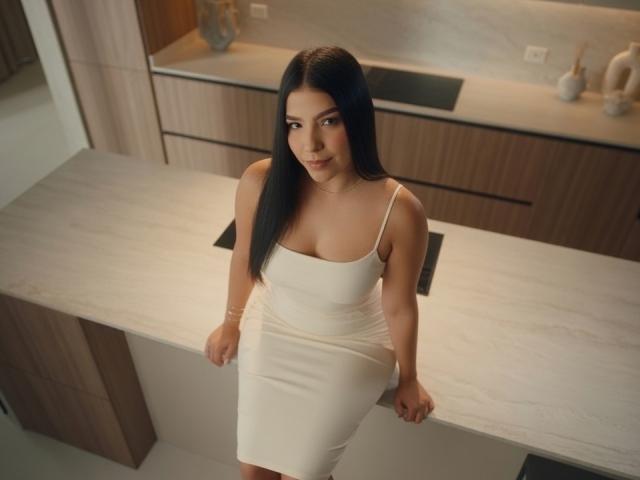 Sofia Delacruz, gekleed in een strakke witte jurk, zit op een aanrecht in de keuken en laat haar kleine borsten en ronde billen zien, met lang zwart haar en een subtiele glimlach..
