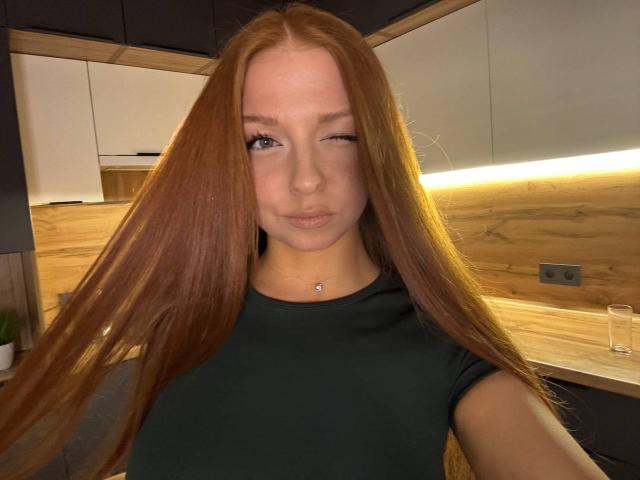 LuanneKadri with long red hair, winking, wearing a black shirt, small silver necklace, and a cute, neutral expression, in a modern kitchen with wooden accents.