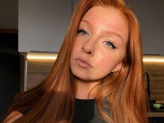 LuanneKadri has long red hair, green eyes, and a slight pout, wearing a black shirt, with small hoop earrings, and a smooth, fair-skinned face.