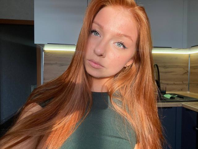 LuanneKadri with long red hair, green eyes, and pink lips, wearing a green top, looking at the camera with a slightly tilted head, in a well-lit kitchen.