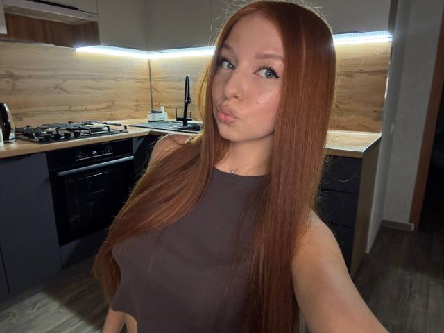 LuanneKadri with long red hair, pouty lips, and blue eyes, wearing a brown sleeveless top, showing her small tits, in a modern kitchen with wooden and dark cabinets.