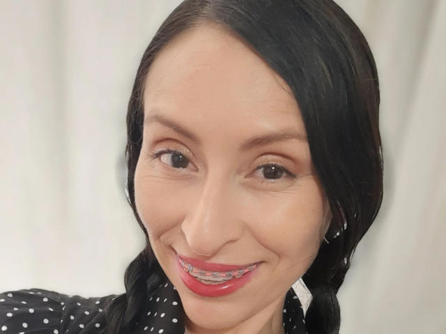 AliceRossiX with dark hair in pigtails, smiling with red lipstick, wearing a black polka dot shirt, and a nose ring, looking cute and sexy.