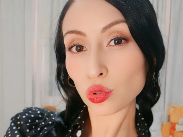 AliceRossiX has glossy red lips, dark wavy hair, brown eyes, and light skin, wearing a black polka dot top with puffed sleeves. Her face is slightly pouty.