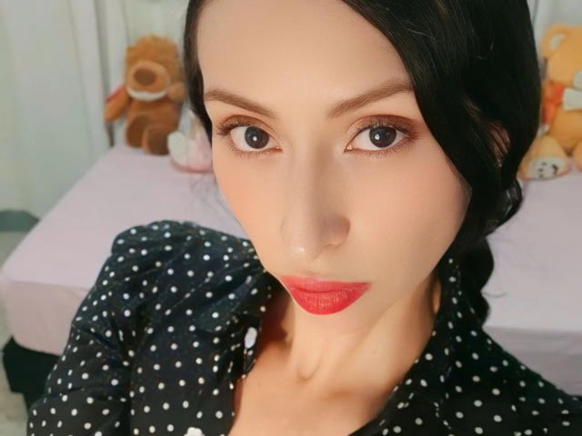 AliceRossiX has pale skin, dark hair in a braid, red lipstick, and wears a black polka dot shirt. Her brown eyes are slightly upturned. She's looking at the camera.
