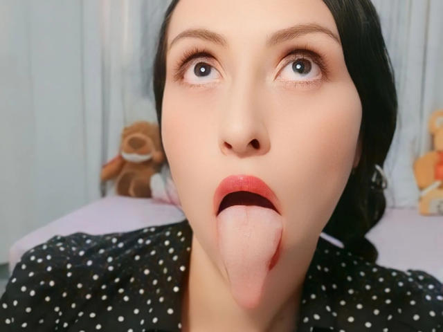 AliceRossiX with her tongue hanging out, eyes wide, wearing a black polka dot shirt, fair skin, black hair, and red lipstick, looking surprised.