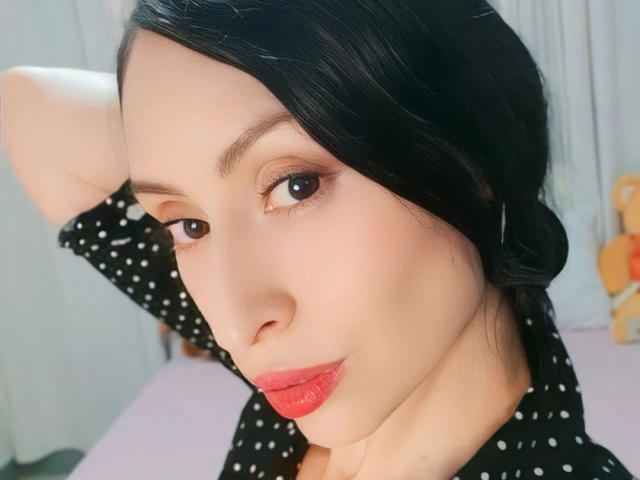 AliceRossiX has pale skin, dark hair in a side braid, red lipstick, brown eyes, and wears a black polka dot shirt. Her arm is raised behind her head.