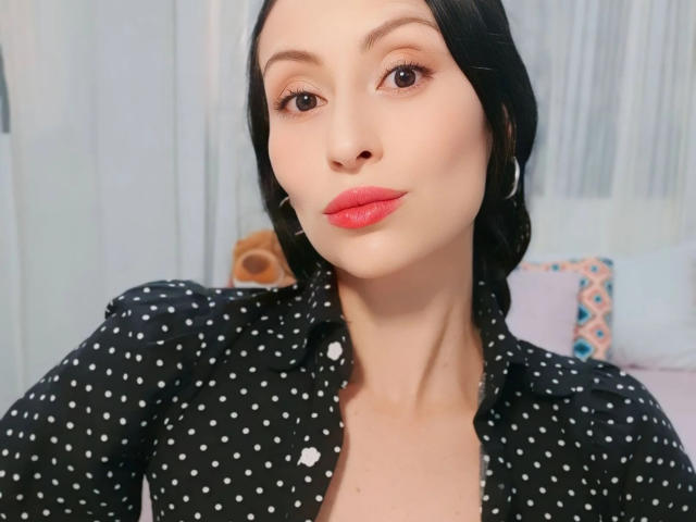 AliceRossiX has pale skin, black hair, red lipstick, and is wearing a black polka dot shirt. She's looking at the camera with a slight smile.