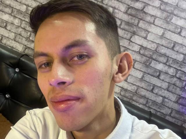 AnderCooper tem um rosto brilhante, levemente avermelhado, com cabelo castanho curto, um pequeno piercing na orelha direita e está vestindo uma camisa branca. Seus lábios estão ligeiramente entreabertos..