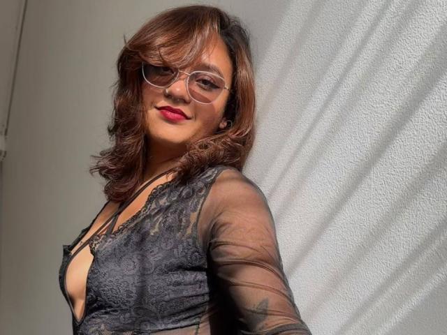 Julie Sharp, con i capelli castano ondulati, rossetto rosso e occhiali, indossa una camicetta trasparente in pizzo nero con un profondo scollo, e sorride alla fotocamera..