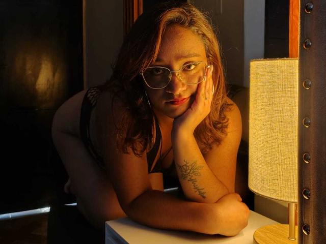 JulieSharp, usando óculos, apoiada em uma mesa, lingerie de renda preta, tatuagem no braço direito, cabelo castanho ondulado, luminária amarela, quarto escuro, olhando para a câmera, mão no rosto..