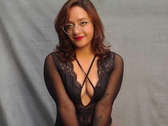 JulieSharp, con la pelle di colore marrone medio, i capelli ricci rossastri, gli occhiali, il rossetto rosso e l'intimo di pizzo nero che mette in mostra il seno e il décolleté, è in piedi contro un tessuto grigio..