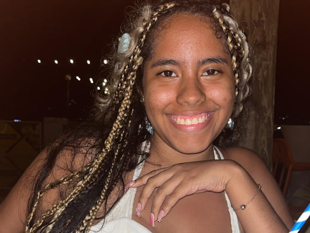 SiennaTaylor with curly blonde and black braids, smiling, tan skin, wearing a white top, showing off pink nails, and a silver bracelet, standing by a wooden pillar.