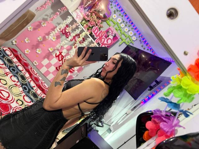MiaBrown69, con lunghi capelli neri, tatuaggi sul braccio, indossa un abito nero senza schienale, sta scattando un selfie allo specchio, tenendo un telefono, con un sedere sexy e formoso..
