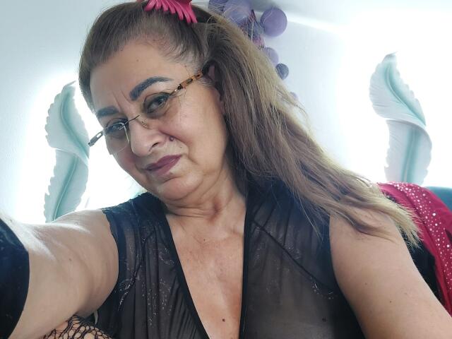 KarlaHartley, middle-aged with glasses, brown hair in a ponytail, pink flower, black sheer top, showing small tits, smiling, light skin, slightly wrinkled face, purple lipstick, purple and white background.
