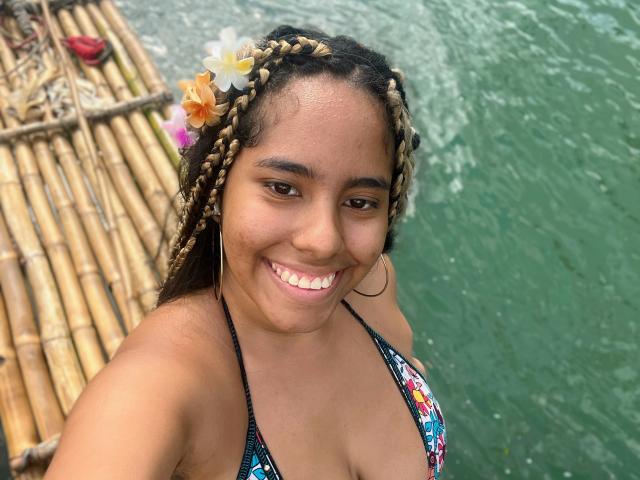 SiennaTaylor with braided hair, flower in hair, wearing a colorful bikini top, smiling, green water background, bamboo raft, hoop earrings, showing her tits, cute, brown skin.