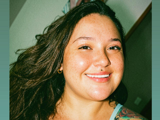 Colette Bessette, con capelli castano scuro ondulati, viso lentigginoso, piercing al naso, sorridente, con un tatuaggio colorato sulla spalla, bordatura verde-blu attorno all'immagine..