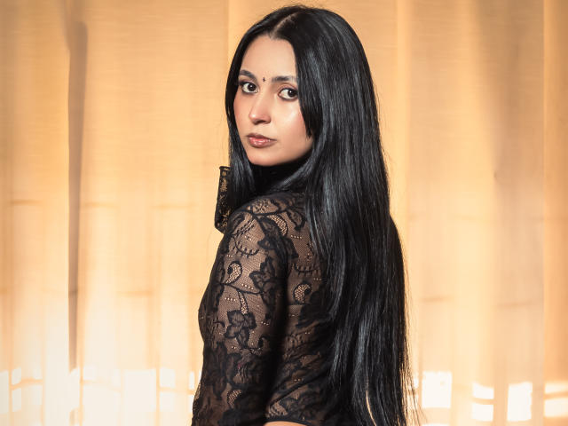 GabyMathew with long black hair, looking over her shoulder, wearing a black lace top, showing her back and subtle makeup, soft light on her face.