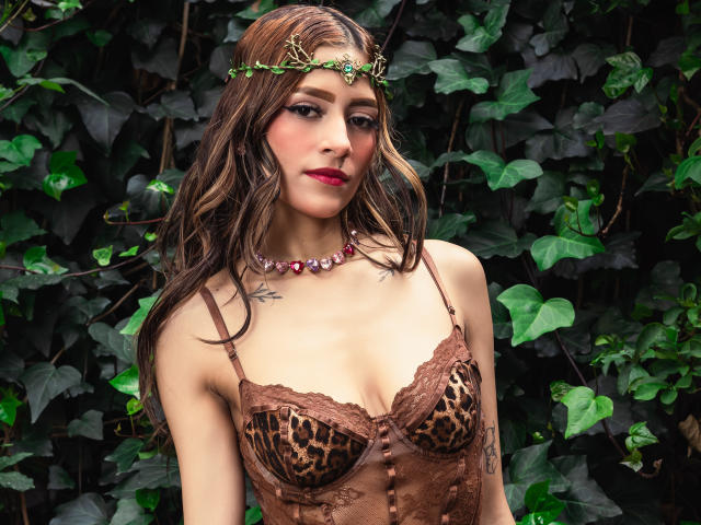 AfroditaWild with brown wavy hair, wearing a leopard print lace bra, green vine crown, pink gem necklace, and red lipstick, has small tits and a tattoo on her right chest.
