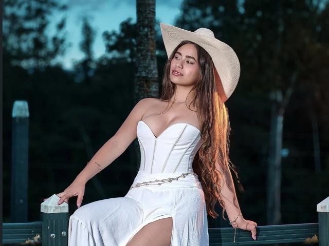 Noribelle, con cabello largo y castaño, vestido blanco sin tirantes y sombrero de vaquera, sentada en un poste de madera, con las manos sobre las rodillas, mostrando sus botas blancas y un cinturón de plata..