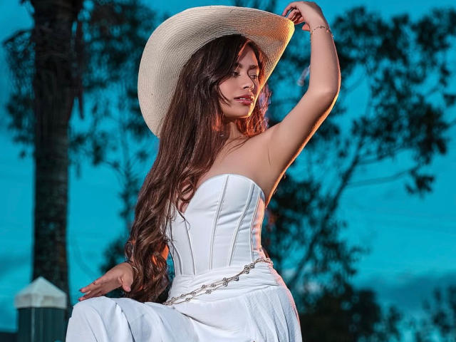 Noribelle, com cabelos castanhos longos, vestindo um vestido branco sem alças e um chapéu de sol, segurando-o com uma mão, luz da noite azulada, mostrando seus seios e nádegas, árvore ao fundo..