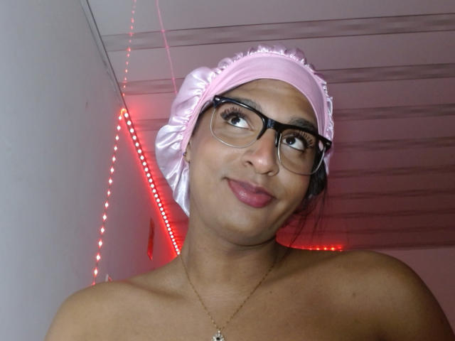 IdiiaLatina with glasses, pink headscarf, and necklace, topless, showing tits, smiling, red LED lights in background, looking up.