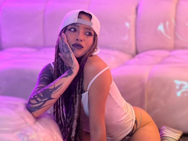 LucyDolly, avec de longues tresses, un débardeur blanc et une casquette à l'envers, montrant son bras tatoué, du rouge à lèvres foncé, et regardant sur le côté, assise sur un canapé blanc sous une lumière rose-violette..