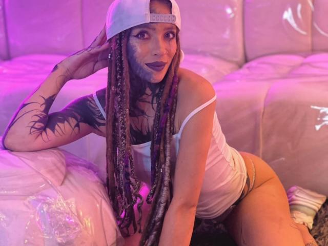 LucyDolly, heiß, mit langen geflochtenen Haaren, weißer Mütze, schwarzem Lippenstift, weißem Tanktop und Unterwäsche, tätowiertem Arm, sitzend auf einer weißen Plastikcouch, mit violetter und pinker Beleuchtung..