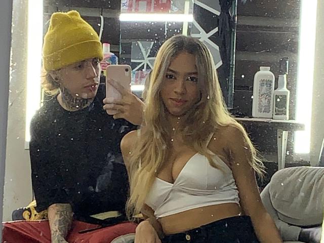 NickySixNine, com cabelos loiros longos, um top branco que mostra seus seios, está ao lado de um cara tatuado com um gorro amarelo, tirando uma selfie no espelho..