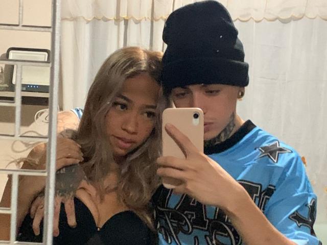 NickySixNine, con lunghi capelli biondi, reggiseno nero e seno di media grandezza, viene tenuta da NickySixNine, che indossa una maglia blu e un berretto nero, mentre si scatta un selfie allo specchio..