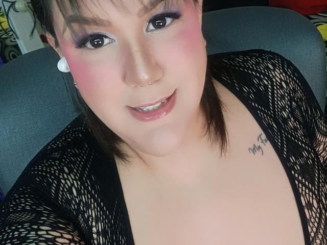 GiiannaMorrys with pink blush, dark eyeliner, nose ring, white earrings, black mesh top, small tattoo on right shoulder, smiling with glossy lips, brown eyes, short brown hair.