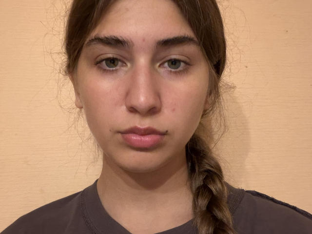 JameStueber a un visage sérieux, des yeux marron clair et une queue de cheval marron tressée. Elle porte une chemise gris foncé avec des détails subtils au niveau du col..