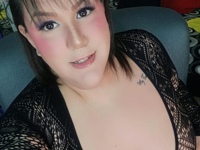 GiiannaMorrys has pink blush, dark eyeliner, and a nose ring, wearing a black fishnet top, showing her tits, with a tattoo on her right chest. She's smiling.