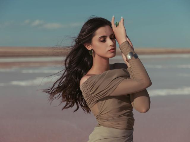 JasmineDay with long dark hair, wearing a tan off-shoulder top, raising her right arm, wearing green earrings and a silver bracelet, standing in a flat, pinkish landscape.