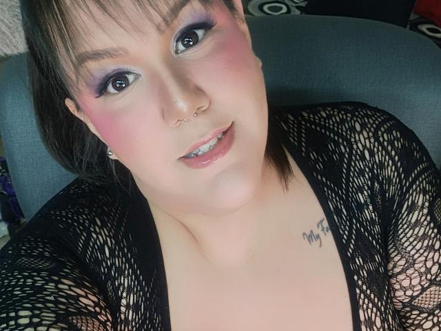 GiiannaMorrys with pink blush, purple eyeshadow, nose ring, and black fishnet top, showing a tattoo on her chest, smiling with slightly parted glossy lips.