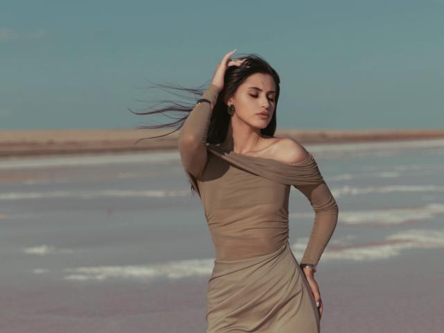 JasmineDay with long black hair, tan off-shoulder dress, hand in hair, eyes closed, standing on beach, wearing bracelet, dress ripples, soft sunlight, calm expression, gentle breeze.
