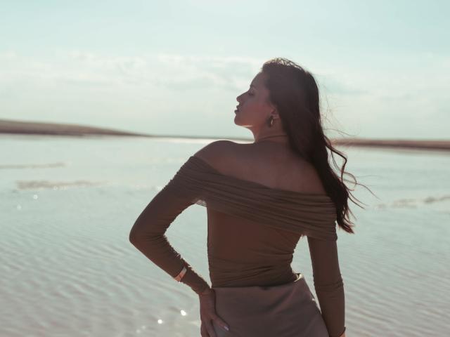 JasmineDay with long brown hair, brown off-shoulder top, and gray skirt, standing by a calm water body, looking back with a slight smile, sunlight in her face.