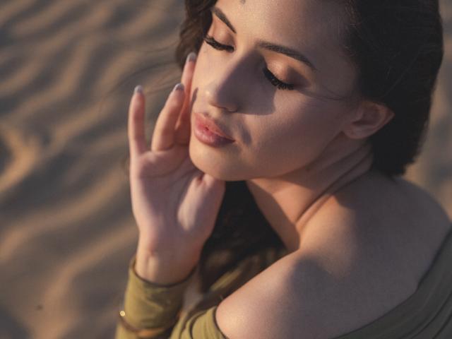 Jasmine, les yeux fermés, la main touchant sa joue, haut vert à une épaule, bracelet doré, cheveux bruns ondulés, fond de sable doux, maquillage discret, expression douce, lèvres légèrement entrouvertes..