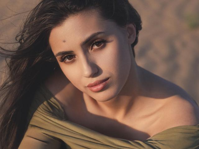 JasmineDay has dark brown eyes, a small mole on her right cheek, and is wearing an olive green off-shoulder top, with her long brown hair flowing to the side.